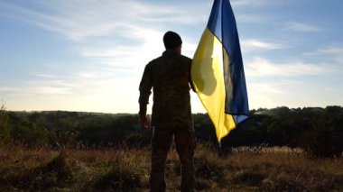 Ukraynalı ordu mensubu, kırsal kesimde ulusal bir pankartıyla tepede duruyor. Rus saldırısına karşı zafer sembolü olarak Ukrayna bayrağı taşıyan askeri üniformalı bir erkek asker. İstila direnişi.