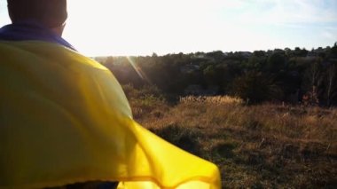 Kamuflaj üniformalı genç adam Ukrayna bayrağı açık havada koşuyor. Rus saldırısına karşı zafer sembolü olarak mavi-sarı bayrak taşıyan Ukraynalı erkek asker. Savaşın sonu..