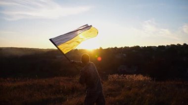 Ukraynalı asker gün batımının arka planına ulusal bayrak sallıyor. Genç erkek asker kırsal kesimde bayrak kaldırdı. Rus saldırısına karşı zafer. Rusya ve Ukrayna arasındaki savaşın sonu..