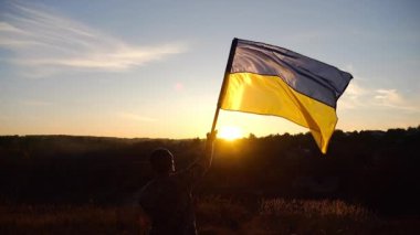 Askeri üniformalı genç adam arka planda gün batımına karşı Ukrayna bayrağı sallıyor. Erkek Ukraynalı asker kırsal kesimde ulusal bayrağı kaldırdı. Rus saldırısına karşı zafer. Savaşın sonu..