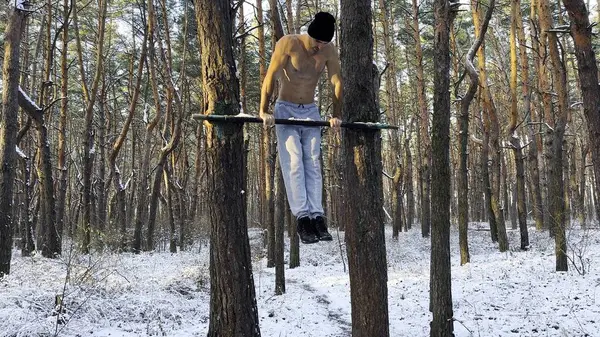 Strong and muscular man doing exercises on horizontal bar at winter forest. Young sportsman muscling up at beautiful nature. Athletic guy training outdoor. Concept of sport and active lifestyle.