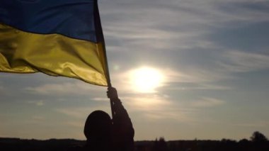 Ukraynalı askerin silueti tepede duruyor ve gün batımına karşı ulusal bayrak sallıyor. Erkek asker, Rus saldırısına karşı zafer sembolü olarak mavi-sarı bayrak kaldırıyor. Bağımsızlık için savaş.
