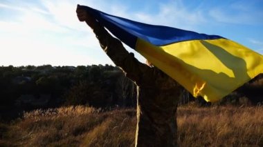 Rus saldırısına karşı kazanılan zaferin şerefine ulusal bayrağı kaldıran Ukraynalı erkek asker. Üniformalı bir asker, Ukrayna bayrağının üzerinde kırsalda dikiliyor. Savaşın sonu..
