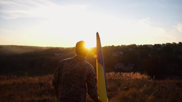 身着迷彩服的年轻人高举蓝黄的旗帜 以庆祝战胜俄国侵略的胜利 乌克兰陆军士兵升起一面飘扬的乌克兰国旗 背景为日落 — 图库视频影像
