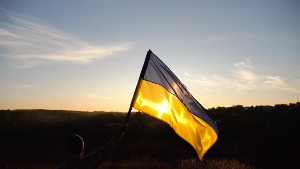 乌克兰陆军士兵升起一面飘扬的乌克兰国旗 背景为日落 身着迷彩服的年轻人高举蓝黄的旗帜 庆祝战胜俄国侵略的胜利 — 图库视频影像