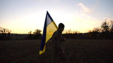 Üniformalı genç erkek asker çayırda Ukrayna bayrağıyla yürüyor. Ukrayna ordusunun askeri gün batımında mavi-sarı pankartıyla sahaya çıkıyor. Rus saldırısına karşı zafer kavramı. Yavaş