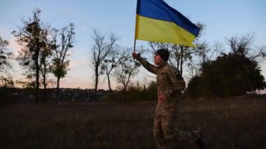Alacakaranlıkta mavi-sarı bayrak taşıyan Ukrayna ordusunun askeri. Çayırda Ukrayna bayrağıyla koşan üniformalı genç bir erkek asker. Rus saldırganlığına karşı zafer.