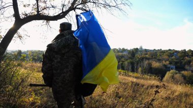 Üniformalı genç erkek asker Ukrayna bayrağıyla kırsalda yürüyor. Ukrayna ordusunun askeri açık havada mavi-sarı bayrak sallıyor. Rus saldırısına karşı zafer kavramı