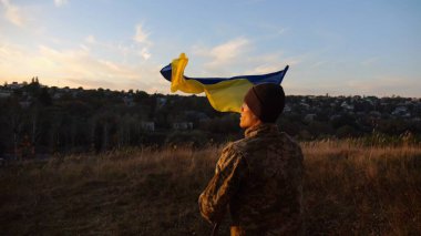 Askeri üniformalı genç adam arka planda güzel günbatımına karşı Ukrayna bayrağı sallıyor. Erkek Ukraynalı asker kırsal kesimde ulusal bayrağı kaldırdı. Rus saldırısına karşı zafer.