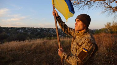 Askeri üniformalı genç adam arka planda güzel günbatımına karşı Ukrayna bayrağı sallıyor. Erkek Ukraynalı asker kırsal kesimde ulusal bayrağı kaldırdı. Rus saldırısına karşı zafer.