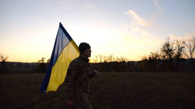 Üniformalı genç erkek asker çayırda Ukrayna bayrağıyla yürüyor. Ukrayna ordusunun askeri gün batımında mavi-sarı pankartıyla sahaya çıkıyor. Rus saldırısına karşı zafer kavramı. Yavaş