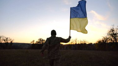 Alacakaranlıkta mavi-sarı bayrak taşıyan Ukrayna ordusunun askeri. Çayırda Ukrayna bayrağıyla koşan üniformalı genç bir erkek asker. Rus saldırganlığına karşı zafer.