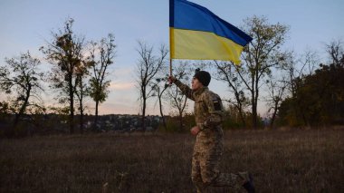 Alacakaranlıkta mavi-sarı bayrak taşıyan Ukrayna ordusunun askeri. Çayırda Ukrayna bayrağıyla koşan üniformalı genç bir erkek asker. Rus saldırganlığına karşı zafer.