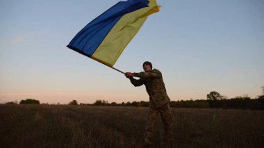 Genç erkek ordu üniformalı Ukrayna bayrağıyla alacakaranlıkta çayırda koşuyor. Ukrayna ordusunun askeri Rus saldırısına karşı zafer şerefine mavi-sarı bayrak sallıyor. Savaşın sonu. Yavaş çekim