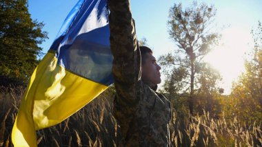 Üniformalı bir asker, Ukrayna bayrağının üzerinde kırsalda dikiliyor. Rus saldırısına karşı kazanılan zaferin şerefine ulusal bayrağı kaldıran Ukraynalı erkek asker. Savaşın sonu.