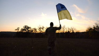Alacakaranlıkta mavi-sarı bayrak taşıyan Ukrayna ordusunun askeri. Çayırda Ukrayna bayrağıyla koşan üniformalı genç bir erkek asker. Rus saldırganlığına karşı zafer.