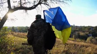 Üniformalı genç erkek asker Ukrayna bayrağıyla kırsalda yürüyor. Ukrayna ordusunun askeri açık havada mavi-sarı bayrak sallıyor. Rus saldırısına karşı zafer kavramı.