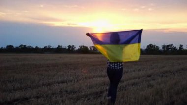 Arpa çayırında yürüyüp Ukrayna bayrağını dalgalandıran mutlu bir kadın. Arka planda güzel bir günbatımında mavi-sarı pankartıyla Ukraynalı bir kadın. Savaşın sonu..
