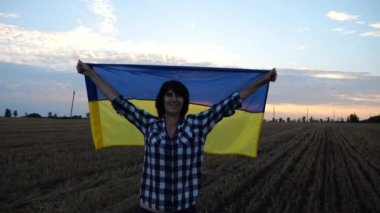 Mutlu kadın gün batımında mavi-sarı bir pankartıyla buğday tarlasında duran kameraya bakıyor. Ukraynalı gülümseyen kadın Ukrayna bayrağının üzerinde gündoğumunda arpa çayırında poz veriyor.