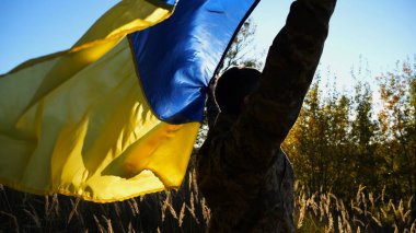 Üniformalı bir asker, Ukrayna bayrağının üzerinde kırsalda dikiliyor. Rus saldırısına karşı kazanılan zaferin şerefine ulusal bayrağı kaldıran Ukraynalı erkek asker. Savaşın sonu.