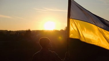 Erkek asker, güzel günbatımının arka planında Ukrayna bayrağıyla ayakta duruyor. Rus saldırısına karşı kazanılan zaferin şerefine mavi-sarı bayrak taşıyan Ukraynalı erkek asker..