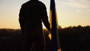 Erkek Ukraynalı asker, güzel günbatımının arka planında ulusal bayrağıyla duruyor. Rus saldırısına karşı zafer sembolü olarak Ukrayna bayrağı taşıyan kamuflaj üniformalı genç adam..