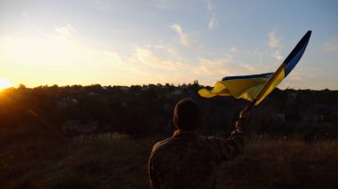 Askeri üniformalı genç adam arka planda güzel günbatımına karşı Ukrayna bayrağı sallıyor. Erkek Ukraynalı asker kırsal kesimde ulusal bayrağı kaldırdı. Rus saldırısına karşı zafer.