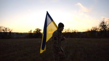 Üniformalı genç erkek asker çayırda Ukrayna bayrağıyla yürüyor. Ukrayna ordusunun askeri gün batımında mavi-sarı pankartıyla sahaya çıkıyor. Rus saldırısına karşı zafer kavramı. Yavaş