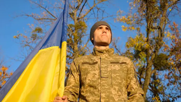 Ukrainian army man stands with national banner at countryside against blue sky. Male soldier in military uniform with Ukraine flag as symbol of victory against russian aggression. Invasion resistance