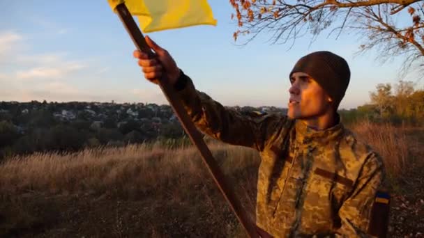 ウクライナの国旗を日没の丘の頂上に振るウクライナ軍の兵士 軍服の若者は田舎で国旗を掲揚した ロシア侵略に対する勝利 戦争の終わり — ストック動画