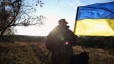 Üniformalı genç erkek asker Ukrayna bayrağıyla kırsalda yürüyor. Ukrayna ordusunun askeri açık havada mavi-sarı bayrak sallıyor. Rus saldırısına karşı zafer kavramı