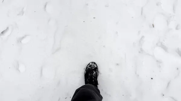 Point of view to male foot stepping on snowy path at winter park. Legs of young man in sneakers kicks up white snow going at forest. Guy walking at nature