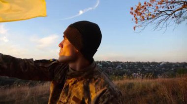 Askeri üniformalı genç adam arka planda gün batımına karşı Ukrayna bayrağı sallıyor. Erkek Ukraynalı asker kırsal kesimde ulusal bayrağı kaldırdı. Rus saldırısına karşı zafer. Savaşın sonu..