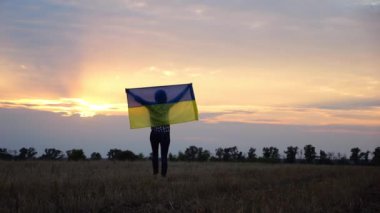 Ukraynalı bir kadın, kafasının üstünde Ukrayna bayrağıyla buğday tarlasında güzel günbatımının arka planına doğru gidiyor. Rus saldırısına karşı zafer sembolü olarak ulusal bayrak taşıyan kadın