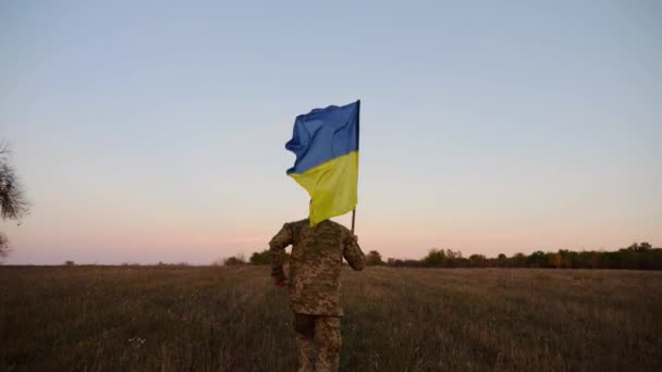 軍服を着た若い男性が ウクライナの国旗を掲げてジョギングをしている ウクライナ陸軍の兵士は 夕暮れにフィールド上に青い黄色の旗を掲げて走った ロシア侵略に対する勝利の概念 — ストック動画