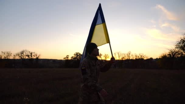 ウクライナ軍の兵士は 日没時にフィールド上に青色の旗を掲げました 制服を着た若い男性は ウクライナの国旗を掲げて歩いている ロシア侵略に対する勝利の概念 — ストック動画