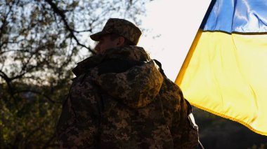 Üniformalı genç erkek asker Ukrayna bayrağıyla kırsalda yürüyor. Ukrayna ordusunun askeri açık havada mavi-sarı bayrak sallıyor. Rus saldırısına karşı zafer kavramı