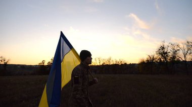 Üniformalı genç erkek asker çayırda Ukrayna bayrağıyla yürüyor. Ukrayna ordusunun askeri gün batımında mavi-sarı pankartıyla sahaya çıkıyor. Rus saldırısına karşı zafer kavramı. Yavaş