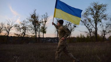 Alacakaranlıkta mavi-sarı bayrak taşıyan Ukrayna ordusunun askeri. Çayırda Ukrayna bayrağıyla koşan üniformalı genç bir erkek asker. Rus saldırganlığına karşı zafer.