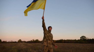 Genç erkek ordu üniformalı Ukrayna bayrağıyla alacakaranlıkta çayırda koşuyor. Ukrayna ordusunun askeri Rus saldırısına karşı zafer şerefine mavi-sarı bayrak sallıyor. Savaşın sonu. Yavaş çekim