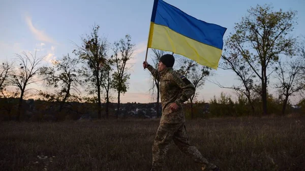 Soldier of ukrainian army running with raised blue-yellow banner on field at dusk. Young male military in uniform jogging with flag of Ukraine at meadow. Victory against russian aggression concept.