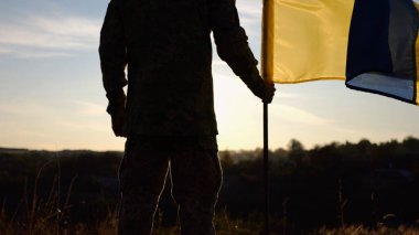 Ukraynalı asker gün batımında kırsal bölgede ulusal bayrağıyla ayakta duruyor. Rus saldırısına karşı zafer sembolü olarak Ukrayna bayrağı taşıyan askeri üniformalı bir erkek asker. İstila direnişi kavramı