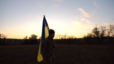 Üniformalı genç erkek asker çayırda Ukrayna bayrağıyla yürüyor. Ukrayna ordusunun askeri gün batımında mavi-sarı pankartıyla sahaya çıkıyor. Rus saldırısına karşı zafer kavramı. Yavaş