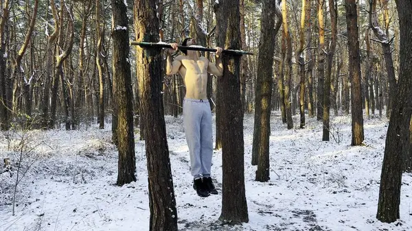 Strong and muscular man exercising on horizontal bar at winter nature. Handsome guy doing pull ups at beautiful forest. Young sportsman training outdoor. Concept of sport and active lifestyle. Slow