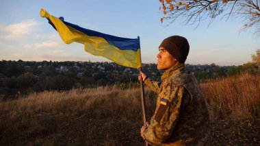 Askeri üniformalı genç adam arka planda güzel günbatımına karşı Ukrayna bayrağı sallıyor. Erkek Ukraynalı asker kırsal kesimde ulusal bayrağı kaldırdı. Rus saldırısına karşı zafer.