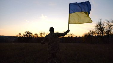 Alacakaranlıkta mavi-sarı bayrak taşıyan Ukrayna ordusunun askeri. Çayırda Ukrayna bayrağıyla koşan üniformalı genç bir erkek asker. Rus saldırganlığına karşı zafer.