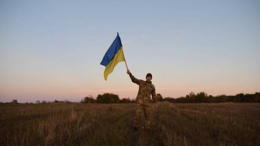 Genç erkek ordu üniformalı Ukrayna bayrağıyla alacakaranlıkta çayırda koşuyor. Ukrayna ordusunun askeri Rus saldırısına karşı zafer şerefine mavi-sarı bayrak sallıyor. Savaşın sonu. Yavaş çekim