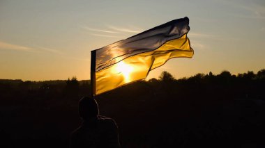 Askeri üniformalı genç adam arka planda güzel günbatımına karşı Ukrayna bayrağı sallıyor. Erkek Ukraynalı asker kırsal kesimde ulusal bayrağı kaldırdı. Rus saldırısına karşı zafer.