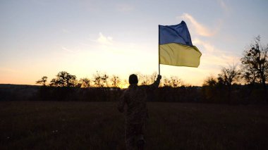 Alacakaranlıkta mavi-sarı bayrak taşıyan Ukrayna ordusunun askeri. Çayırda Ukrayna bayrağıyla koşan üniformalı genç bir erkek asker. Rus saldırganlığına karşı zafer.