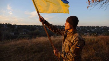 Askeri üniformalı genç adam arka planda güzel günbatımına karşı Ukrayna bayrağı sallıyor. Erkek Ukraynalı asker kırsal kesimde ulusal bayrağı kaldırdı. Rus saldırısına karşı zafer.